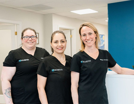 Three women in scrubs stand by a reception desk in a dental office, smiling and ready to assist patients.