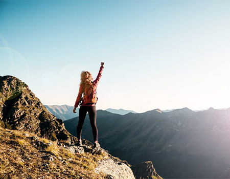 Woman standing on a mountain peak with arms raised in celebration.